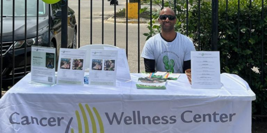 A man wearing sunglasses and a Cancer Wellness Center t-shirt sits behind an information table covered with brochures and flyers. The tablecloth reads "Cancer Wellness Center" and colorful balloons are attached to a fence behind him.