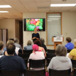 A woman stands at the front of a room giving a presentation to a seated group of adults. A screen behind her displays colorful vegetables and the words “Pancreatic Wellness.”.