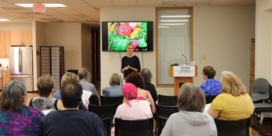 A woman stands at the front of a room giving a presentation to a seated group of adults. A screen behind her displays colorful vegetables and the words “Pancreatic Wellness.”.