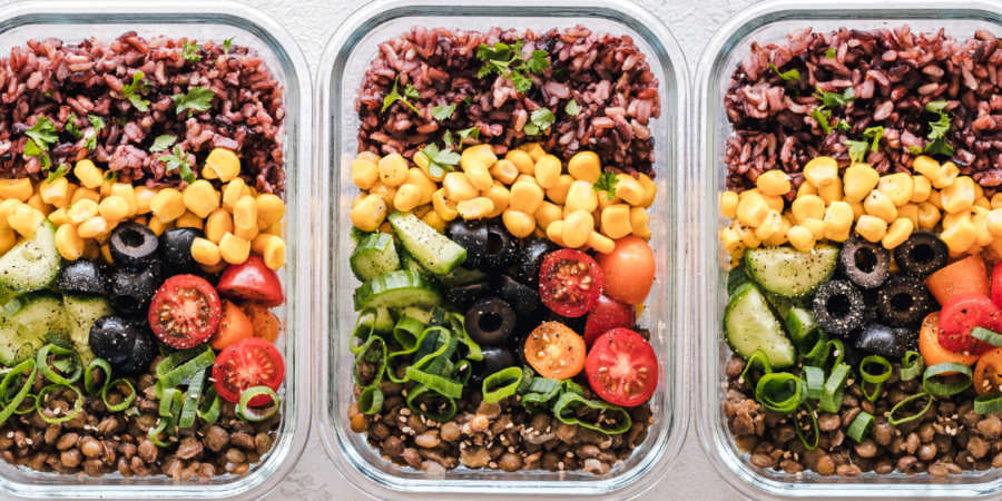 Three glass containers filled with layered meal prep bowls, featuring black rice, corn, cherry tomatoes, cucumber, black olives, lentils, and green onions, arranged neatly on a light-colored surface.