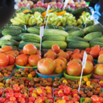 Fresh produce, including tomatoes, cucumbers, peppers, and cherry tomatoes, displayed in rows of baskets and bins at a farmers market. White signs are placed among the colorful fruits and vegetables.
