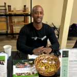 A man sits at a table with brochures, a basket of wrapped candy, and a cup of coffee. He wears a “Cancer Wellness Center” shirt and name tag, smiling at the camera in an indoor setting.
