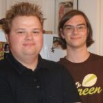 Two young men stand indoors, smiling at the camera. One has short, spiked hair and wears a dark collared shirt; the other has glasses and shoulder-length hair, wearing a brown shirt with green and white lettering.