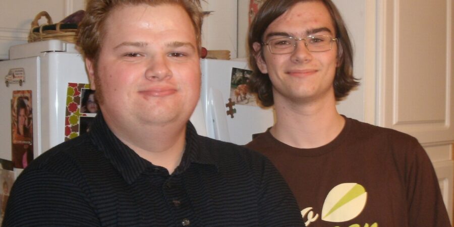 Two young men stand indoors, smiling at the camera. One has short, spiked hair and wears a dark collared shirt; the other has glasses and shoulder-length hair, wearing a brown shirt with green and white lettering.