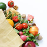 A brown paper bag filled with fresh fruits, vegetables, nuts, and bread, including tomatoes, strawberries, cucumber, radishes, an apple, an orange, a peach, arugula, asparagus, walnuts, an avocado, and a bread roll.