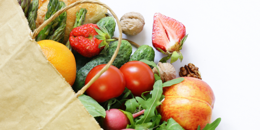A brown paper bag filled with fresh fruits, vegetables, nuts, and bread, including tomatoes, strawberries, cucumber, radishes, an apple, an orange, a peach, arugula, asparagus, walnuts, an avocado, and a bread roll.