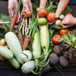 A group of hands reaching toward a variety of fresh vegetables on a wooden table, including zucchini, carrots, corn, tomatoes, onions, lettuce, potatoes, and green beans.