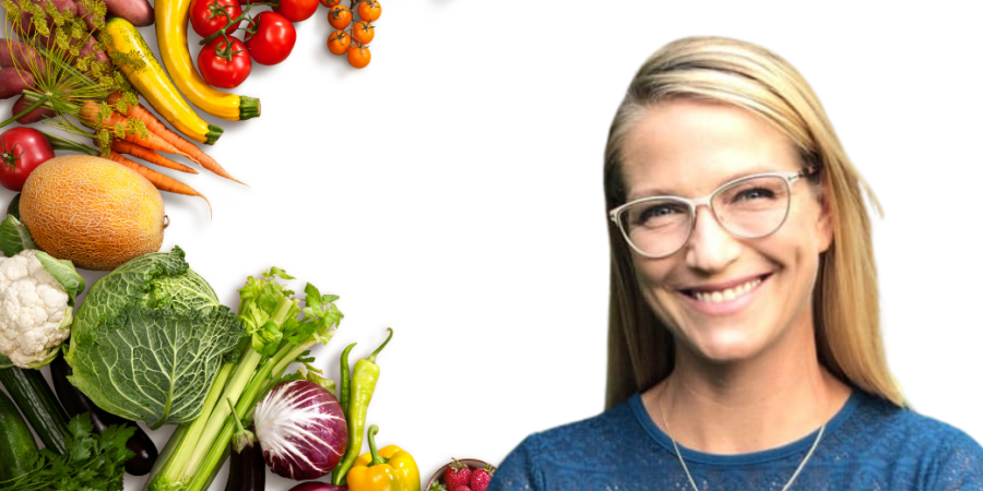 A woman with long blonde hair and glasses smiles in front of a white background, with various fresh vegetables and fruits, like tomatoes, carrots, and melons, arranged along the left side.