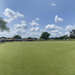 People are standing around the edge of a large golf green under a blue sky with scattered clouds. Several golfers are lining up putts, while spectators watch from the background. Trees border the scene.