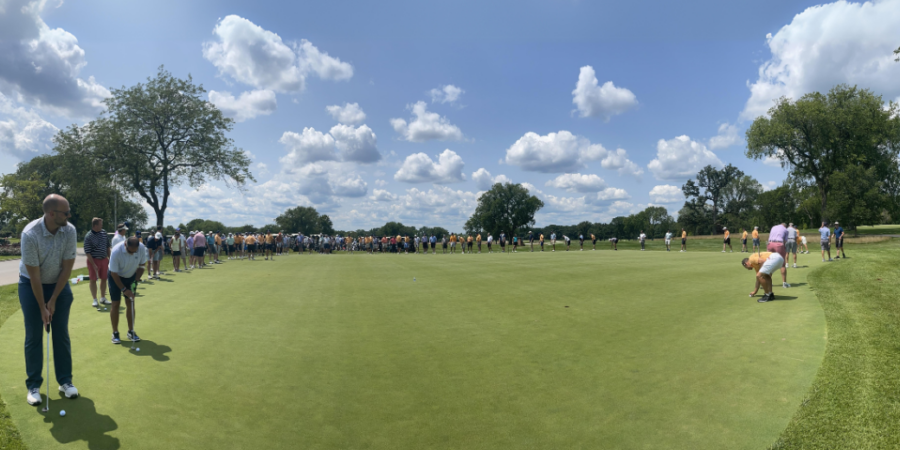 People are standing around the edge of a large golf green under a blue sky with scattered clouds. Several golfers are lining up putts, while spectators watch from the background. Trees border the scene.