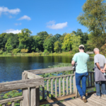 Three people stand on a wooden platform overlooking a pond with lily pads, surrounded by trees under a clear blue sky. The group appears to be enjoying the peaceful, sunny outdoor scenery.