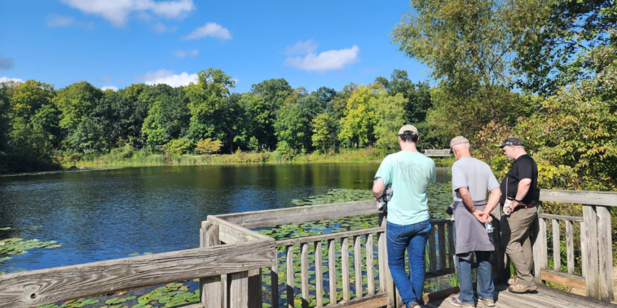 Three people stand on a wooden platform overlooking a pond with lily pads, surrounded by trees under a clear blue sky. The group appears to be enjoying the peaceful, sunny outdoor scenery.