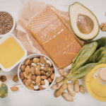 A selection of healthy foods including salmon, avocado, eggs, spinach, broccoli, nuts, seeds, and oil, all arranged on a light wooden surface.
