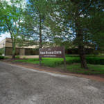 A brown sign reading "Campus Wellness Center" stands in front of a beige brick building surrounded by green trees and shrubs, along a paved driveway under a partly cloudy sky.