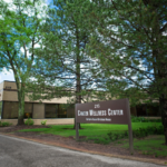 A brown sign reading "Campus Wellness Center" stands in front of a beige brick building surrounded by green trees and shrubs, along a paved driveway under a partly cloudy sky.