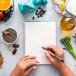 A person writes in a blank notebook surrounded by healthy foods, a scale, dumbbells, and a tape measure, symbolizing nutrition, fitness, and meal planning.