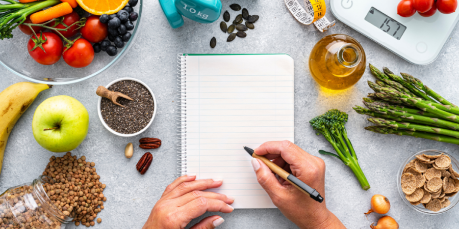 A person writes in a blank notebook surrounded by healthy foods, a scale, dumbbells, and a tape measure, symbolizing nutrition, fitness, and meal planning.