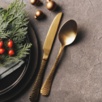 Elegant holiday table setting with a gray napkin, sprigs of greenery, and red berries tied with a ribbon on a dark plate, beside gold-colored knife and spoon on a textured gray surface.