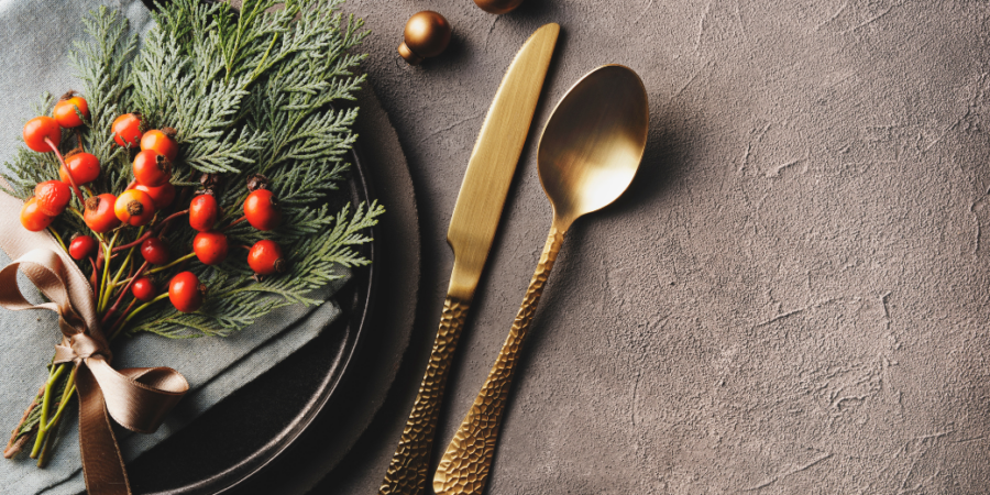 Elegant holiday table setting with a gray napkin, sprigs of greenery, and red berries tied with a ribbon on a dark plate, beside gold-colored knife and spoon on a textured gray surface.