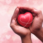 Two hands gently cup a red heart with a white, looping line across it, set against a soft pink background filled with faint heart shapes.