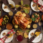 A roasted turkey with apples and orange slices is at the center of a table set for four, with plates, leafy napkins, vegetables, cranberry sauce, pie, and fall leaves as decoration. Glasses of juice complete the autumn feast.