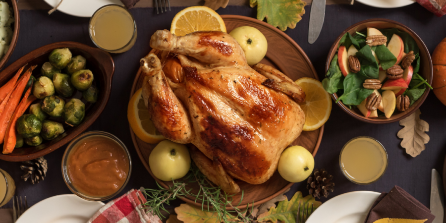 A roasted turkey with apples and orange slices is at the center of a table set for four, with plates, leafy napkins, vegetables, cranberry sauce, pie, and fall leaves as decoration. Glasses of juice complete the autumn feast.