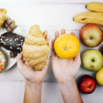 Two hands hold a croissant in the left and an orange in the right, with assorted pastries, bananas, and apples on a white wooden table in the background.