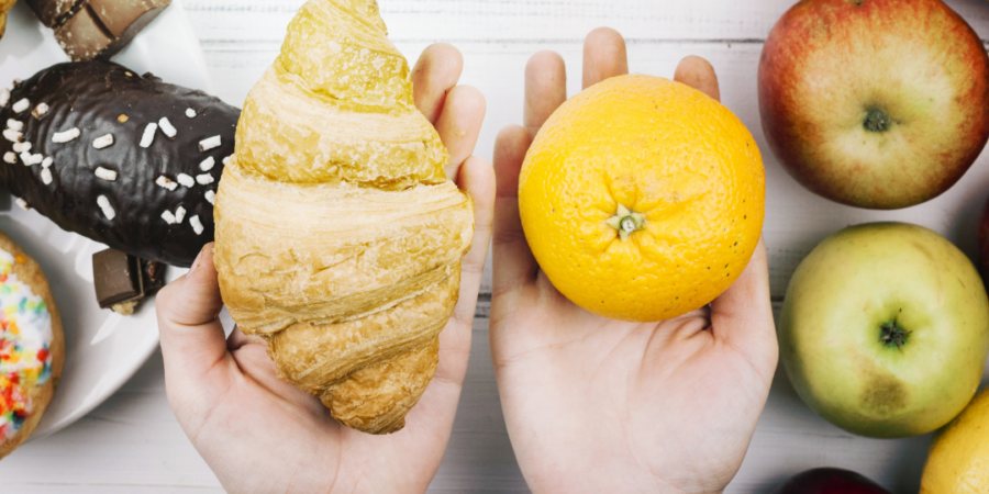 Two hands hold a croissant in the left and an orange in the right, with assorted pastries, bananas, and apples on a white wooden table in the background.