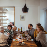 A multi-generational family sits around a dining table sharing a meal, laughing and hugging each other in a warmly lit room. Plates of food, glasses of wine, and a bottle are on the table.