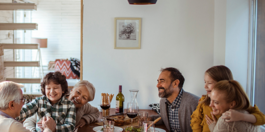 A multi-generational family sits around a dining table sharing a meal, laughing and hugging each other in a warmly lit room. Plates of food, glasses of wine, and a bottle are on the table.