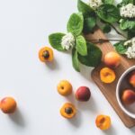 A white bowl with apricots sits on a wooden board next to green leaves and white flowers. Several whole and halved apricots are scattered on a white surface.