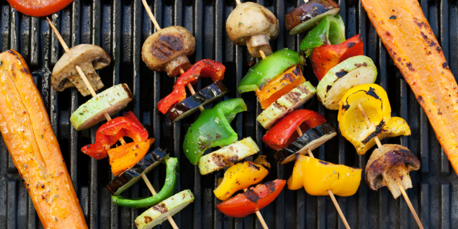 Colorful vegetable skewers with mushrooms, bell peppers, zucchini, and eggplant grilling on a barbecue, alongside long slices of carrot on the grill grates.