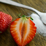 A whole strawberry and a halved strawberry sit on a wooden surface next to a metal spoon filled with white granulated sugar.