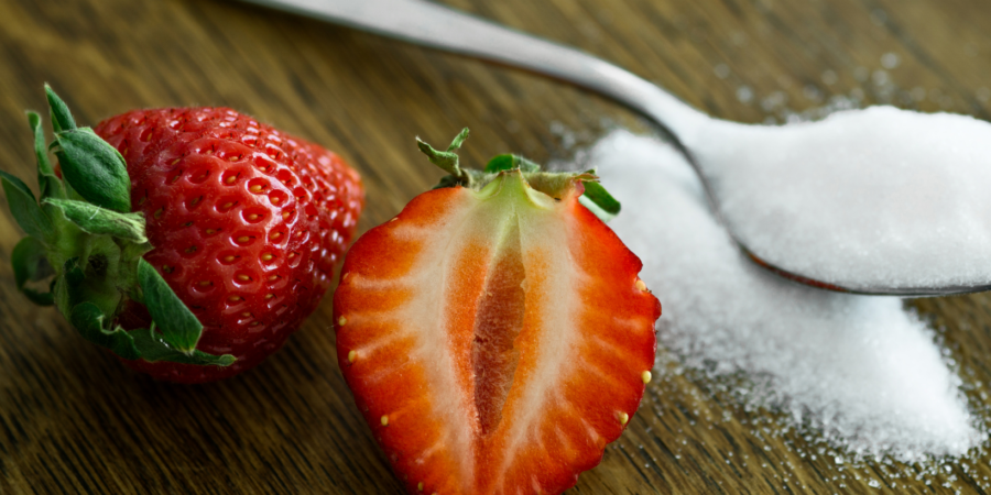 A whole strawberry and a halved strawberry sit on a wooden surface next to a metal spoon filled with white granulated sugar.