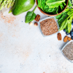 A variety of healthy foods including spinach, avocado, asparagus, blueberries, pecans, celery, flaxseeds, chia seeds, olive oil, and broccoli arranged on a light textured surface.