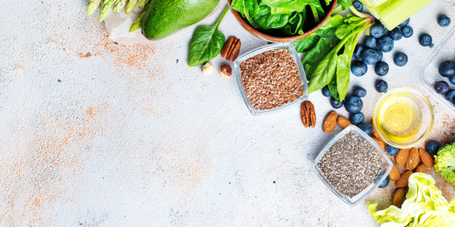 A variety of healthy foods including spinach, avocado, asparagus, blueberries, pecans, celery, flaxseeds, chia seeds, olive oil, and broccoli arranged on a light textured surface.