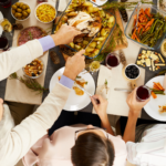 People sharing a festive meal at a table, with dishes including roasted chicken, potatoes, carrots, green beans, cheese, olives, and glasses of wine, viewed from above.