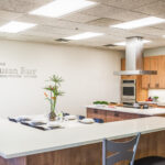 Modern demonstration kitchen with wood cabinets, stainless steel appliances, a vent hood, and a white island counter set with plates and cutlery. A bowl of fruit and a tall plant are on the counter. "Susan Barr Demonstration Kitchen" text on wall.
