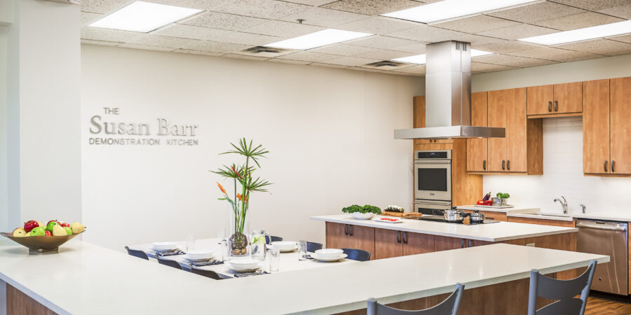Modern demonstration kitchen with wood cabinets, stainless steel appliances, a vent hood, and a white island counter set with plates and cutlery. A bowl of fruit and a tall plant are on the counter. "Susan Barr Demonstration Kitchen" text on wall.