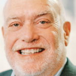 An older man with a bald head and white beard smiles warmly at the camera. He is wearing a suit jacket and a light-colored shirt, with a blurred cityscape background.