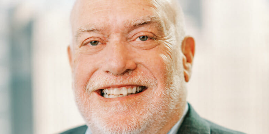An older man with a bald head and white beard smiles warmly at the camera. He is wearing a suit jacket and a light-colored shirt, with a blurred cityscape background.