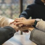 Four people stack their hands together in a gesture of teamwork and unity, wearing casual and business attire. The focus is on their hands, with blurred faces and background.