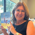 A woman with glasses and shoulder-length hair sits on a couch, smiling and holding up a children's book titled "¡Te quiero, Mari!" with an illustrated cover. Sunlight streams in through a window behind her.