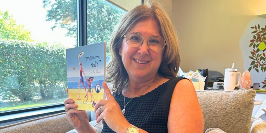A woman with glasses and shoulder-length hair sits on a couch, smiling and holding up a children's book titled "¡Te quiero, Mari!" with an illustrated cover. Sunlight streams in through a window behind her.