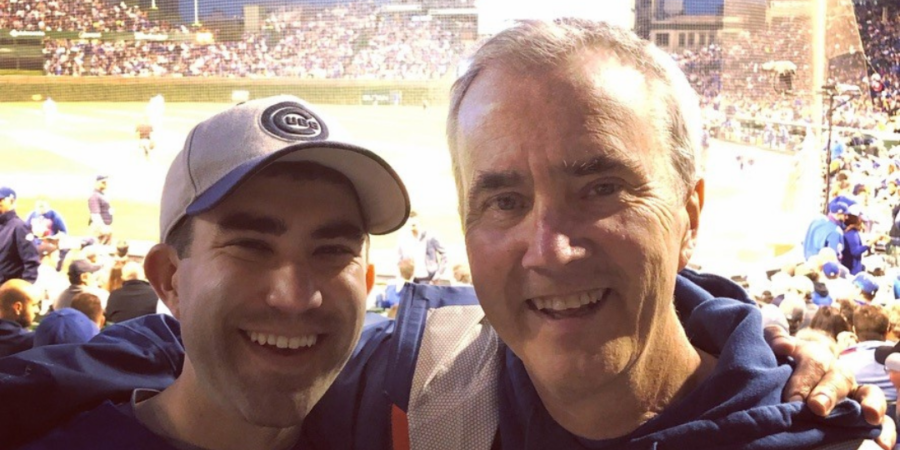 Two men smiling with their arms around each other at a baseball stadium. Both wear blue jackets and baseball caps. The field and a crowd of fans are visible in the background under the evening sky.