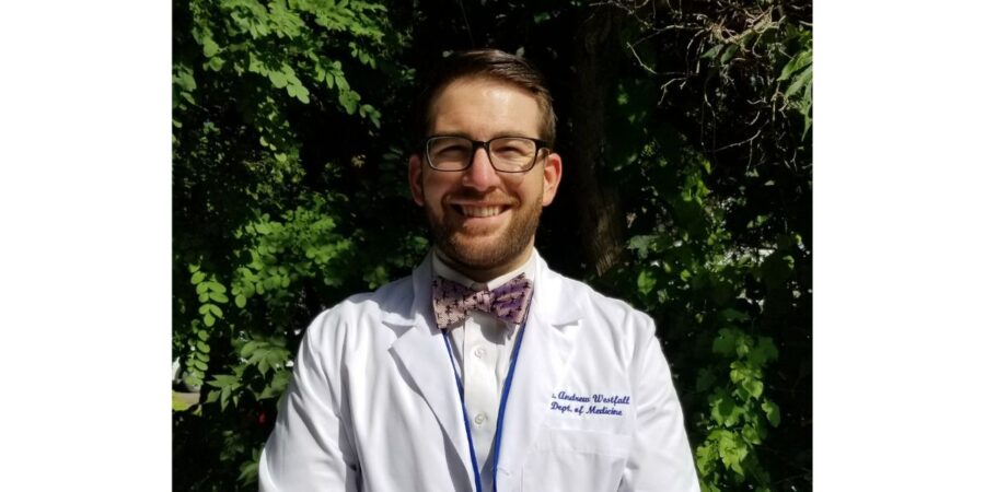 A man wearing glasses, a white lab coat, and a bow tie stands smiling outdoors in front of leafy green trees. The lab coat has text on the chest.