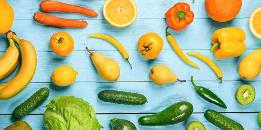 Variety of fresh fruits and vegetables, including bananas, lettuce, carrots, peppers, oranges, apples, kiwi, grapes, cucumbers, and tomatoes, arranged on a bright blue wooden surface.