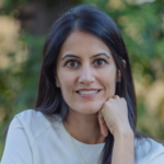 A woman with long dark hair and a white top smiles softly at the camera, resting her chin on her hand. The background is blurred with greenery.