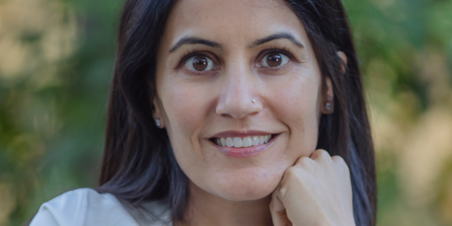 A woman with long dark hair and a white top smiles softly at the camera, resting her chin on her hand. The background is blurred with greenery.