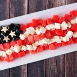 A rectangular platter with blueberries, star-shaped cheese slices, watermelon cubes, and white cheese cubes arranged to resemble the American flag on a wooden background.